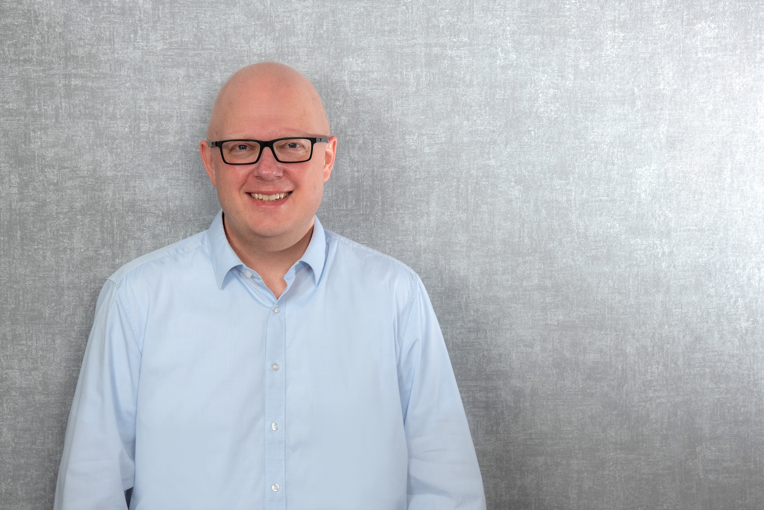Thorsten Jungmann, ein Mann mit kurzen Haaren, dunkler Brille und hellblauem Hemd vor grauem Fotohintergrund