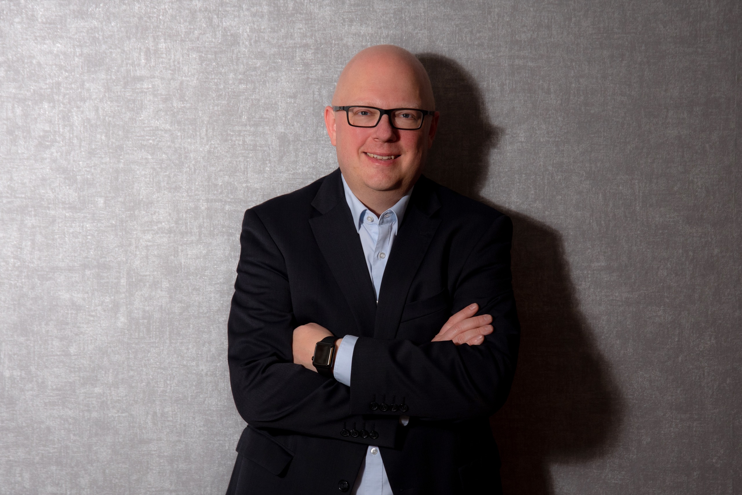 Thorsten Jungmann, ein Mann mit kurzen, hellen Haaren, dunkler Brille, hellblauem Hemd und dunkelblauem Sakko, vor einem grauen Fotohintergrund