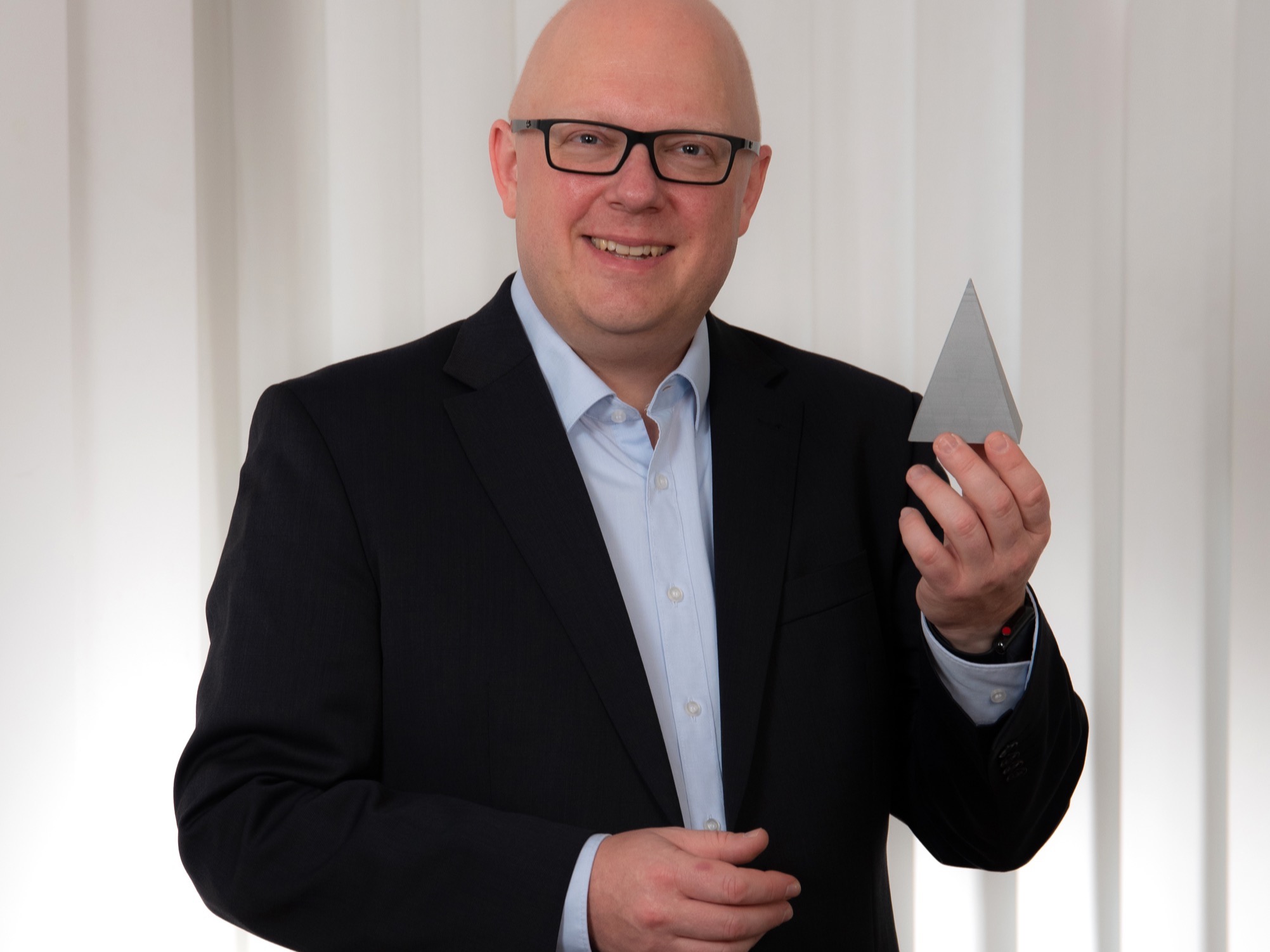 Thorsten Jungmann, ein Mann mit kurzen, hellen Haaren, dunkler Brille, hellblauem Hemd und dunkelblauem Sakko, hält eine Figur in der Hand
